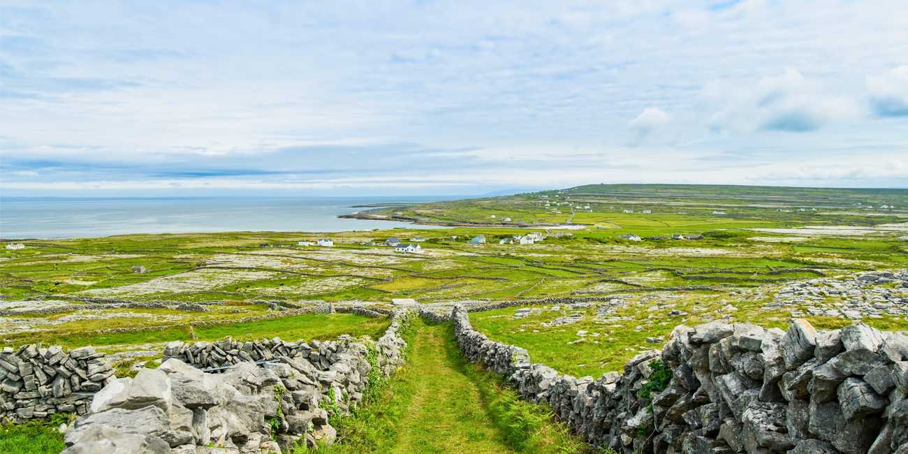 Absorb Ireland's traditional heritage – Discover the timeless landscapes, historic stone walls, and rugged Atlantic coast of the Aran Islands in 2026 A peaceful green grassy path winding between hand-built grey dry-stone walls leading down toward the deep blue Atlantic Ocean on the Aran Islands in 2026